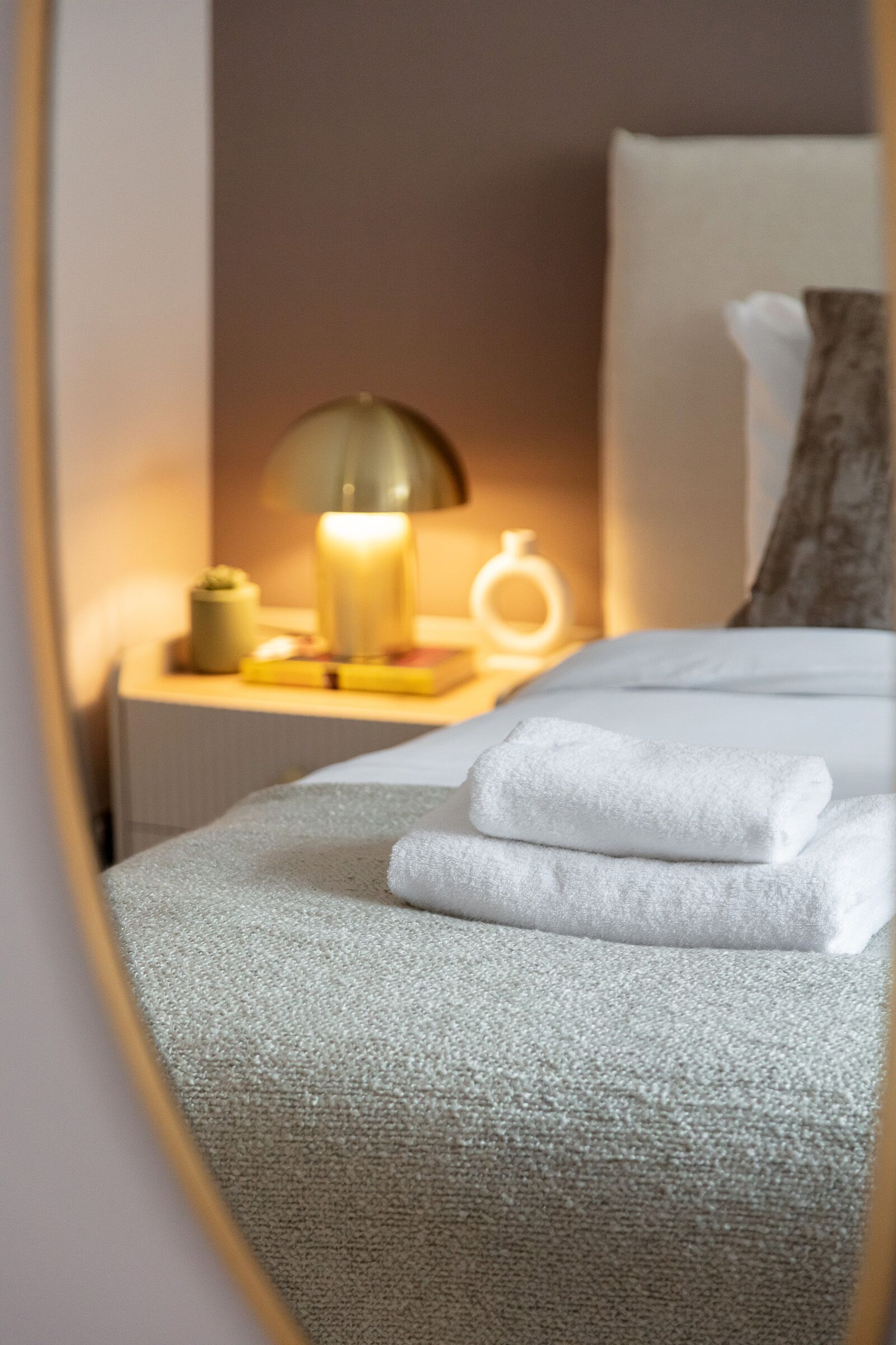 Un lit bien fait, avec du linge de lit blanc et une serviette pliée, une lampe de chevet et une table de chevet sur laquelle se trouvent une bougie et un petit objet décoratif.