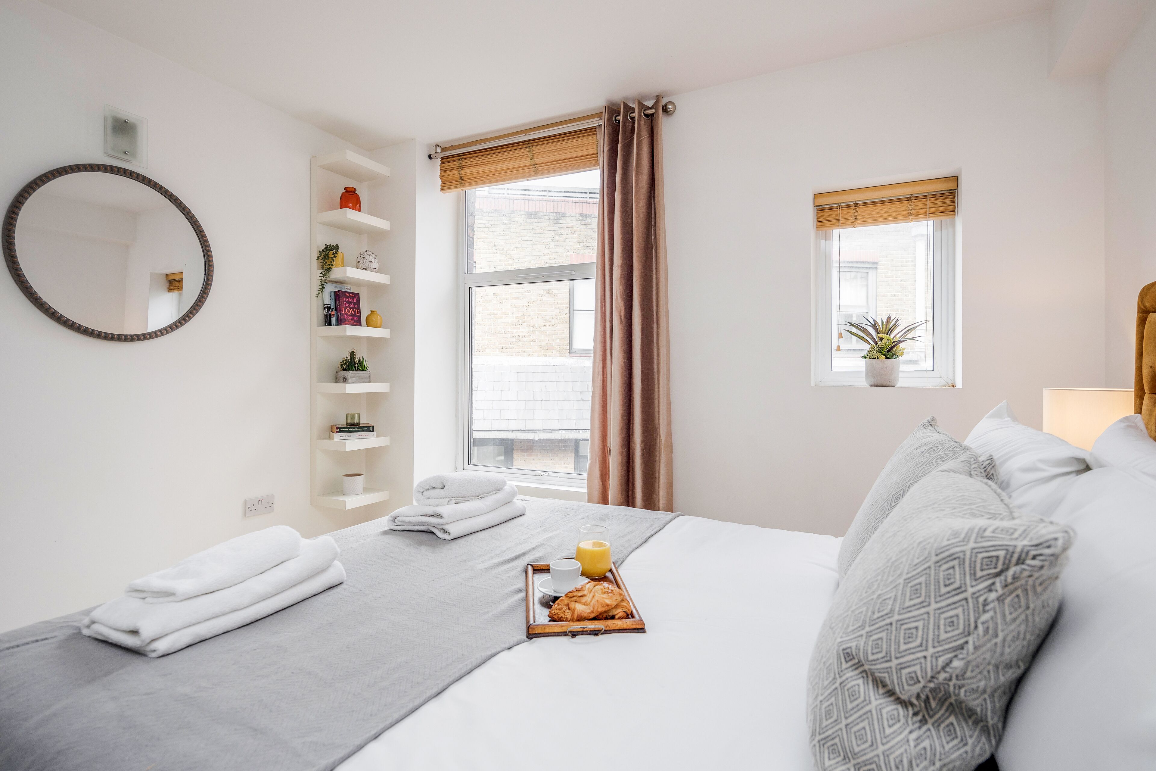 A neatly arranged hotel room with a bed, a bedside table with a tray of breakfast, a window with curtains, a shelf with toiletries, and a mirror on the wall.