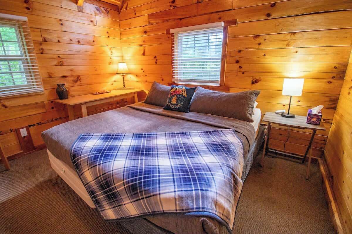 A cozy wooden cabin bedroom with a bed, two bedside tables, and a window with blinds.