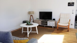 En moderne stue med et fladskærms-tv, et hvidt sofabord, en sofa med mønstrede hynder, en træstol og en lille kaktusplante.