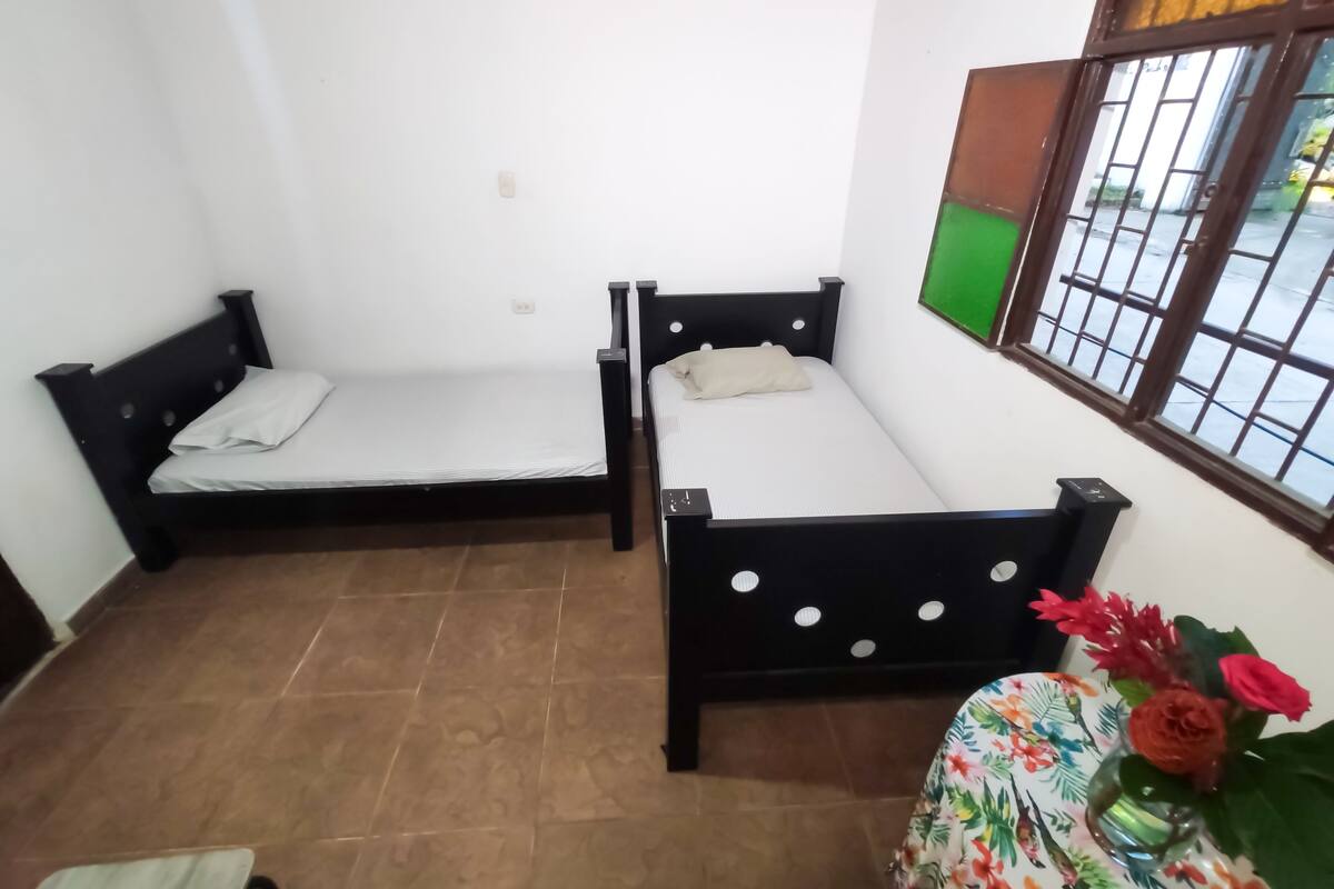 A small room with two single beds, a window with bars, and a table with a floral tablecloth and a vase of flowers.