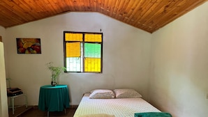 Une chambre avec un plafond en bois, un lit recouvert d’une courtepointe aux couleurs vives, une fenêtre aux vitres jaunes et vertes, et une petite table recouverte d’une nappe verte.