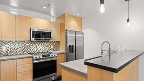 A modern kitchen with wooden cabinets, a black countertop, stainless steel appliances, and a tiled backsplash. - 2 mins 2 Mill Ave + ASU | Pool + EV Charging + GYM (Tempe)