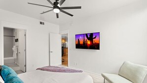 A bedroom with a bed, a chair, a ceiling fan, and a wall-mounted TV displaying a desert sunset. - 2 mins 2 Mill Ave + ASU | Pool + EV Charging + GYM (Tempe)