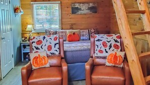 En mysig stuga med en inredd loftvåning som sovplats, en trästege och två läderfåtöljer med kuddar i Halloween-tema.