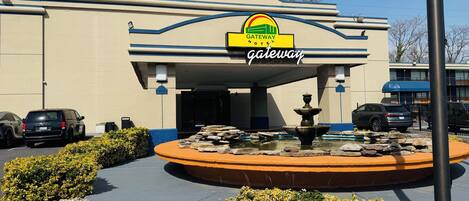 Gateway Hotel com uma fonte à frente, estacionamento e céu azul límpido.