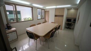 Une salle de conférence avec une longue table et des chaises, un coin cuisine équipé d’un réfrigérateur et d’une machine à café, et une fenêtre donnant sur un paysage verdoyant.