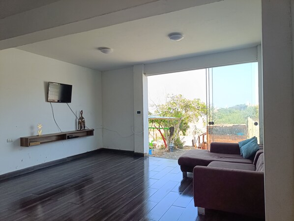 A spacious living room with a dark wooden floor, a mounted flat-screen TV, and a sliding glass door leading to an outdoor area with a view of greenery and a building in the distance.