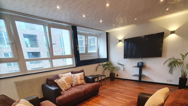 A spacious living room with a large sectional sofa, a flat-screen TV mounted on the wall, and large windows providing natural light. - 3Beds Family Friendly in London with good transport links. Close to Victoria (London)