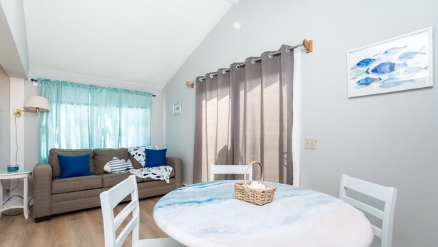 A living room with a round table, white chairs, a sofa with blue cushions, and a window with curtains.