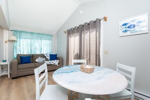 Ein Wohnzimmer mit rundem Tisch, weißen Stühlen, einer Couch mit blauen Kissen und einem Fenster mit Vorhängen.