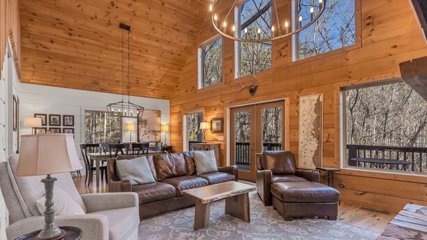 Interior acogedor de una cabaña con techo de madera, sofás de cuero, zona de comedor y ventanas amplias que ofrecen vistas del bosque circundante.
