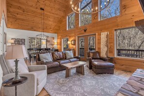 A cozy cabin interior with a wooden ceiling, leather sofas, a dining area, and large windows offering views of the surrounding forest.