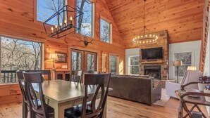 Una cabaña de madera espaciosa con comedor, sala de estar con chimenea de piedra y ventanas amplias con vistas al bosque circundante.