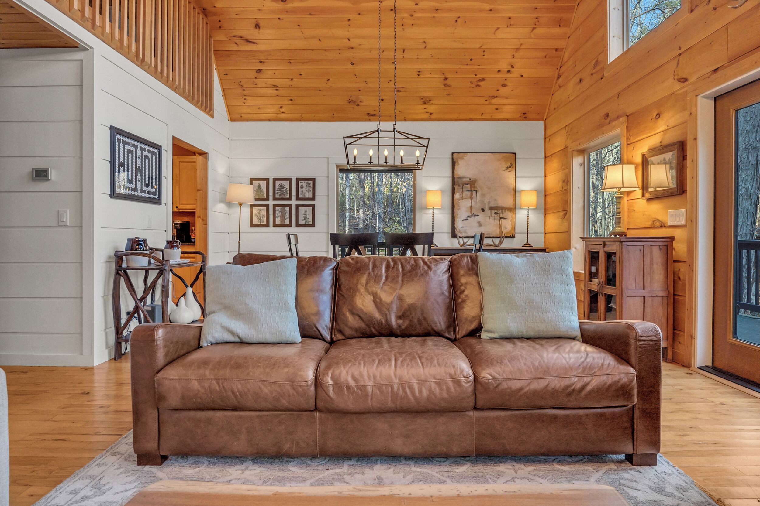 A spacious living room with a brown leather sofa, wooden ceiling, and large windows.