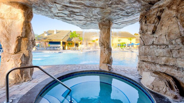 Ein Poolbereich mit einem steinernen Bogen, einem Whirlpool und Blick auf ein Resortgebäude mit Pool und Liegebereich.