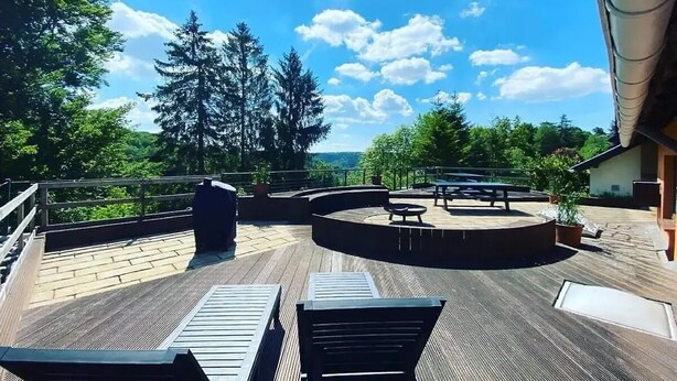 Terraza en la azotea con piso de madera, mobiliario exterior y vistas a árboles y colinas.
