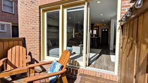 Une terrasse avec des chaises en bois et une petite table, une porte-fenêtre coulissante donnant sur un espace intérieur et un mur en briques.