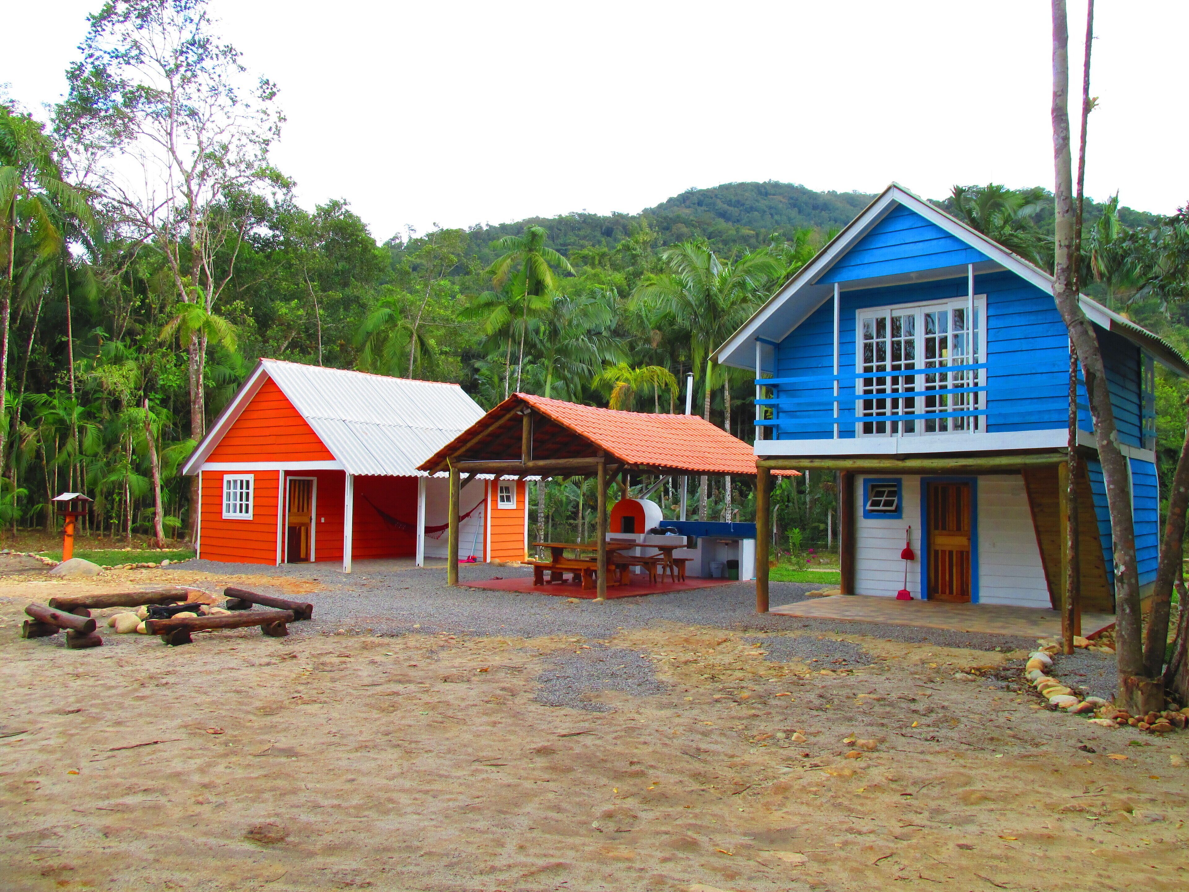 Dos cabañas coloridas con fachadas rojas y azules, rodeadas por un paisaje natural con árboles y una colina al fondo.