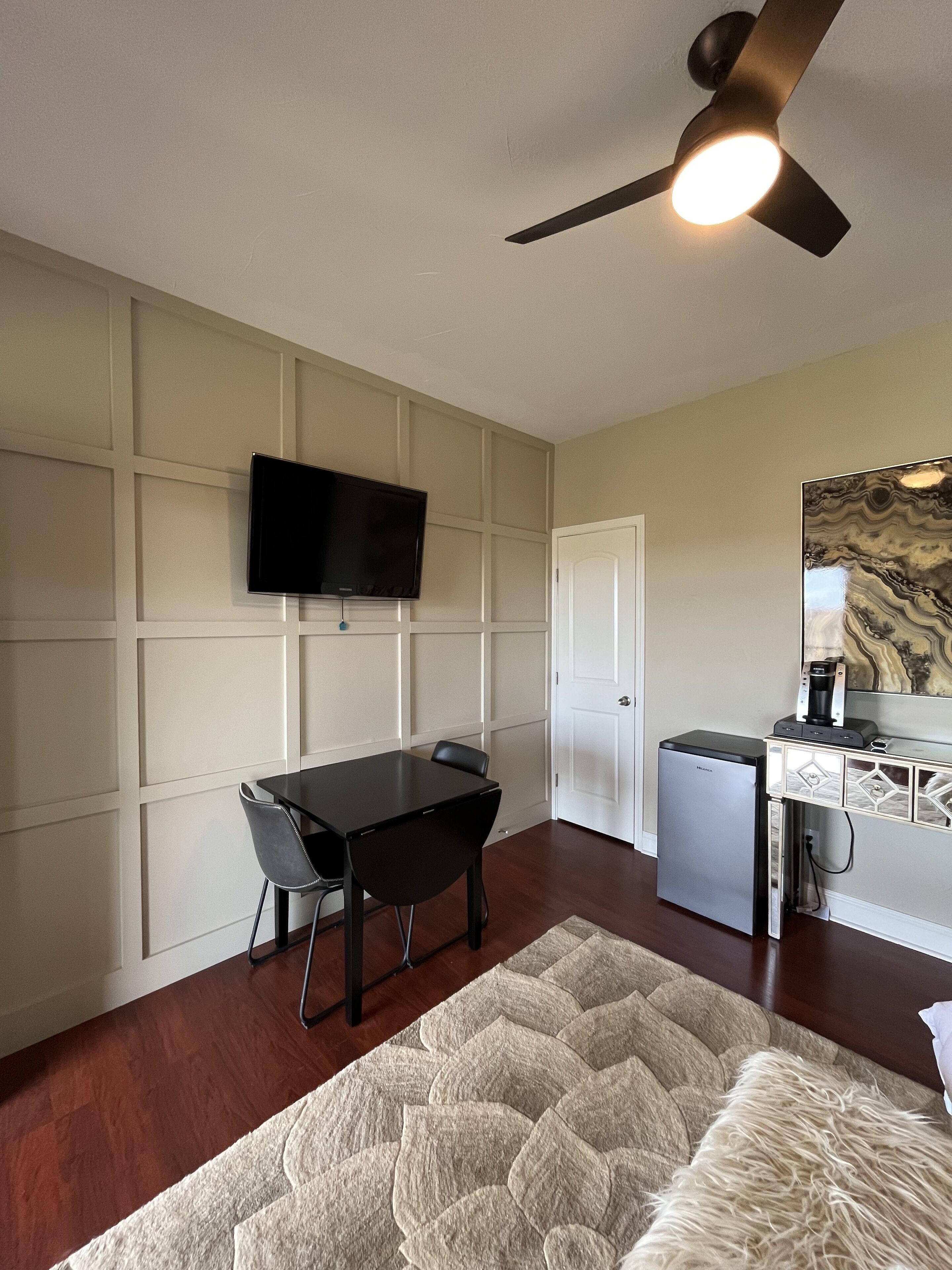 A hotel room with a dining table, chairs, a TV mounted on the wall, a ceiling fan, a refrigerator, and a patterned rug.