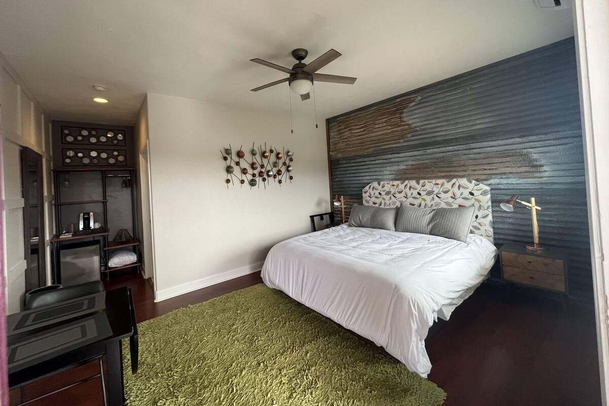 A modern bedroom with a bed, a ceiling fan, a wall art piece, and a nightstand with a lamp.