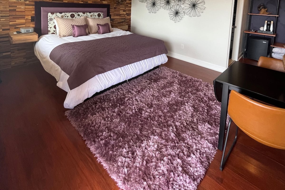 A bedroom with a bed, a ceiling fan, a wooden wall, a decorative wall art, and a small table with a chair.