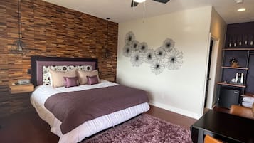 A bedroom with a bed, a ceiling fan, a wooden wall, a decorative wall art, and a small table with a chair.
