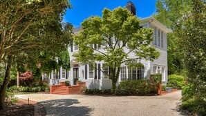 Een wit huis met twee verdiepingen, een bakstenen ingang, omgeven door weelderige beplanting en een grindpad.