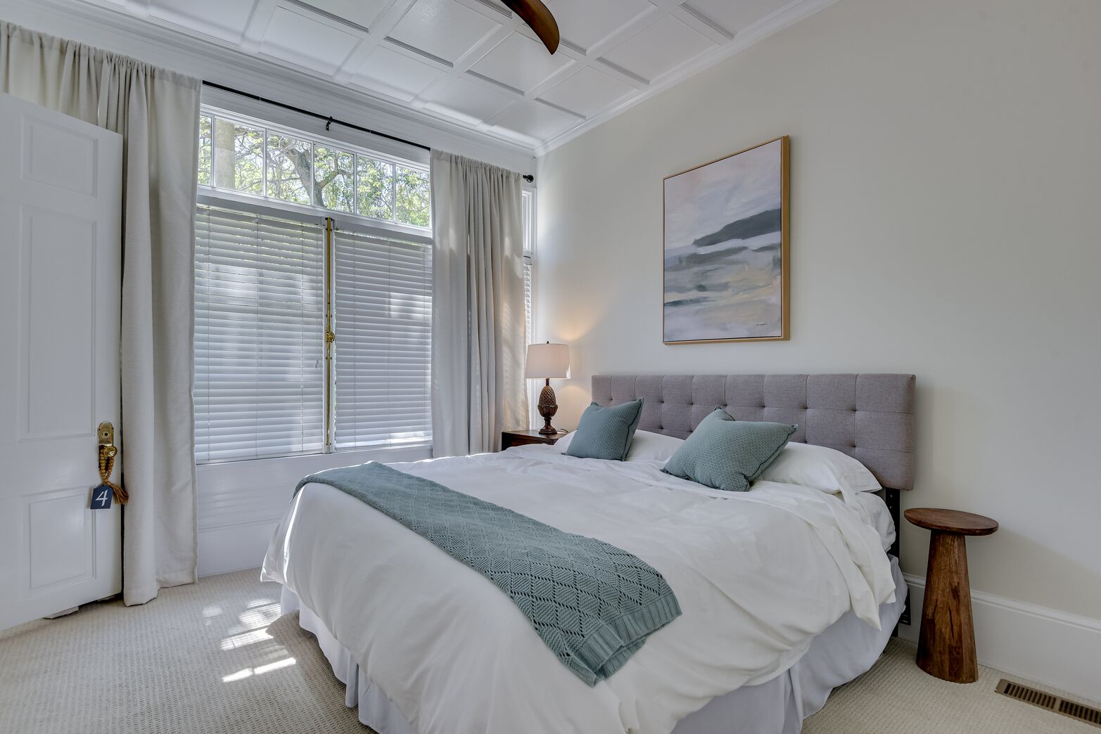 Une chambre avec un grand lit, une table de chevet avec une lampe, une fenêtre avec des stores et un tableau au mur.
