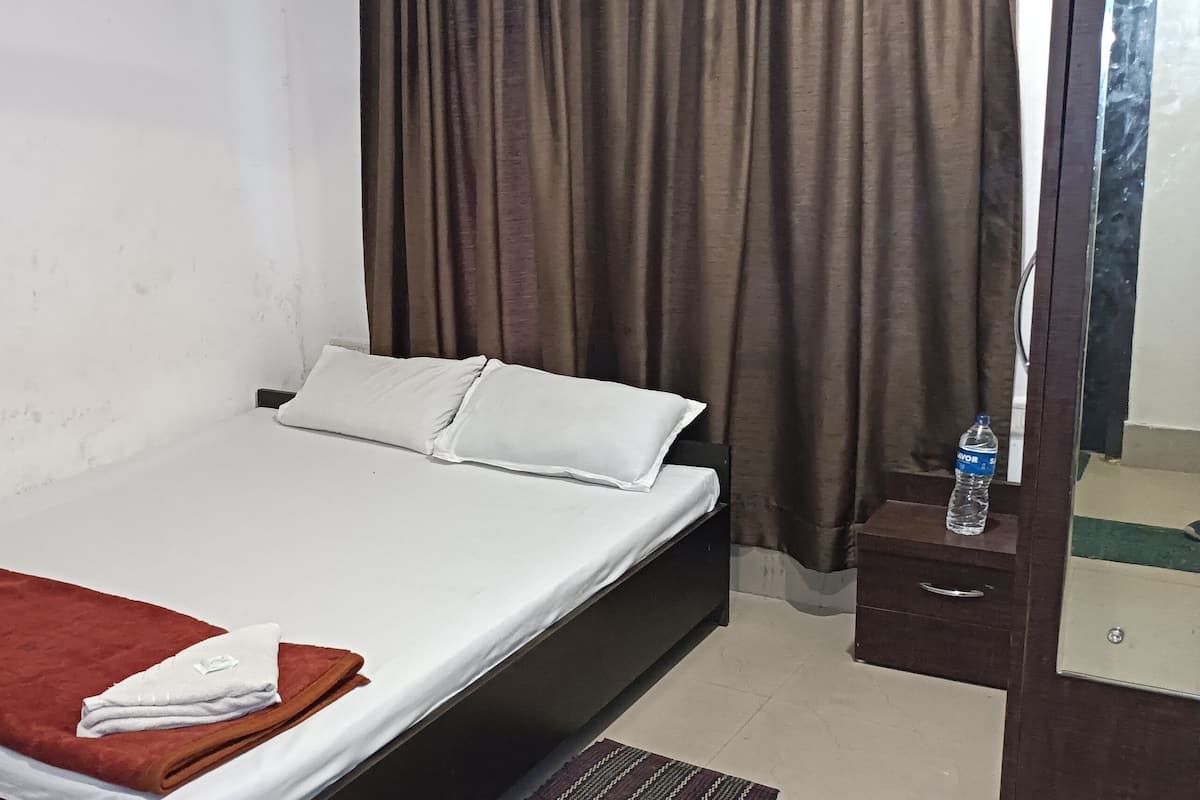 A single bed with a white mattress and two pillows, a brown blanket, and a small bedside table with a water bottle.