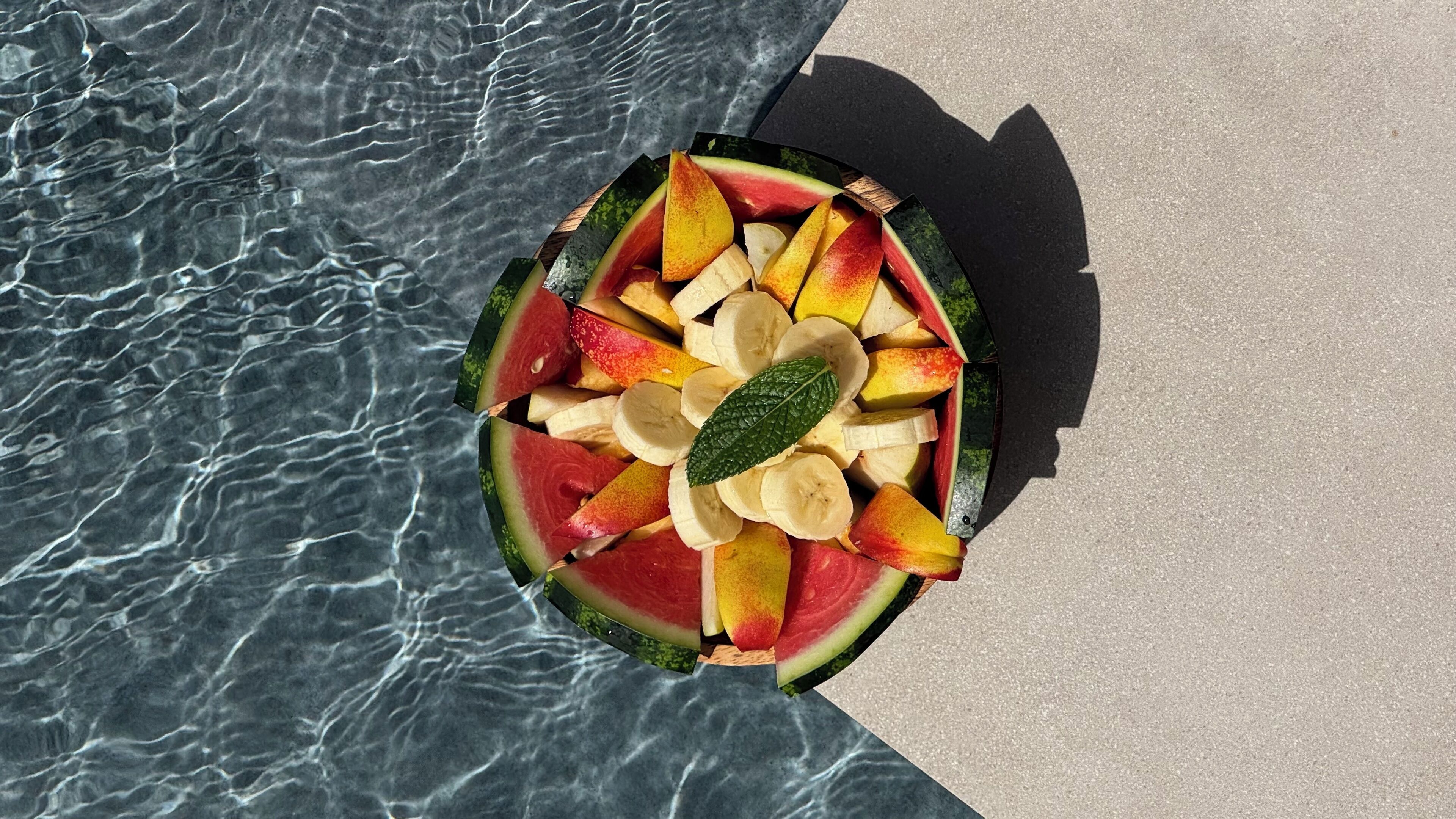 Une pastèque évidée, remplie de tranches de pommes et surmontée d’une banane, est posée au bord de la piscine.