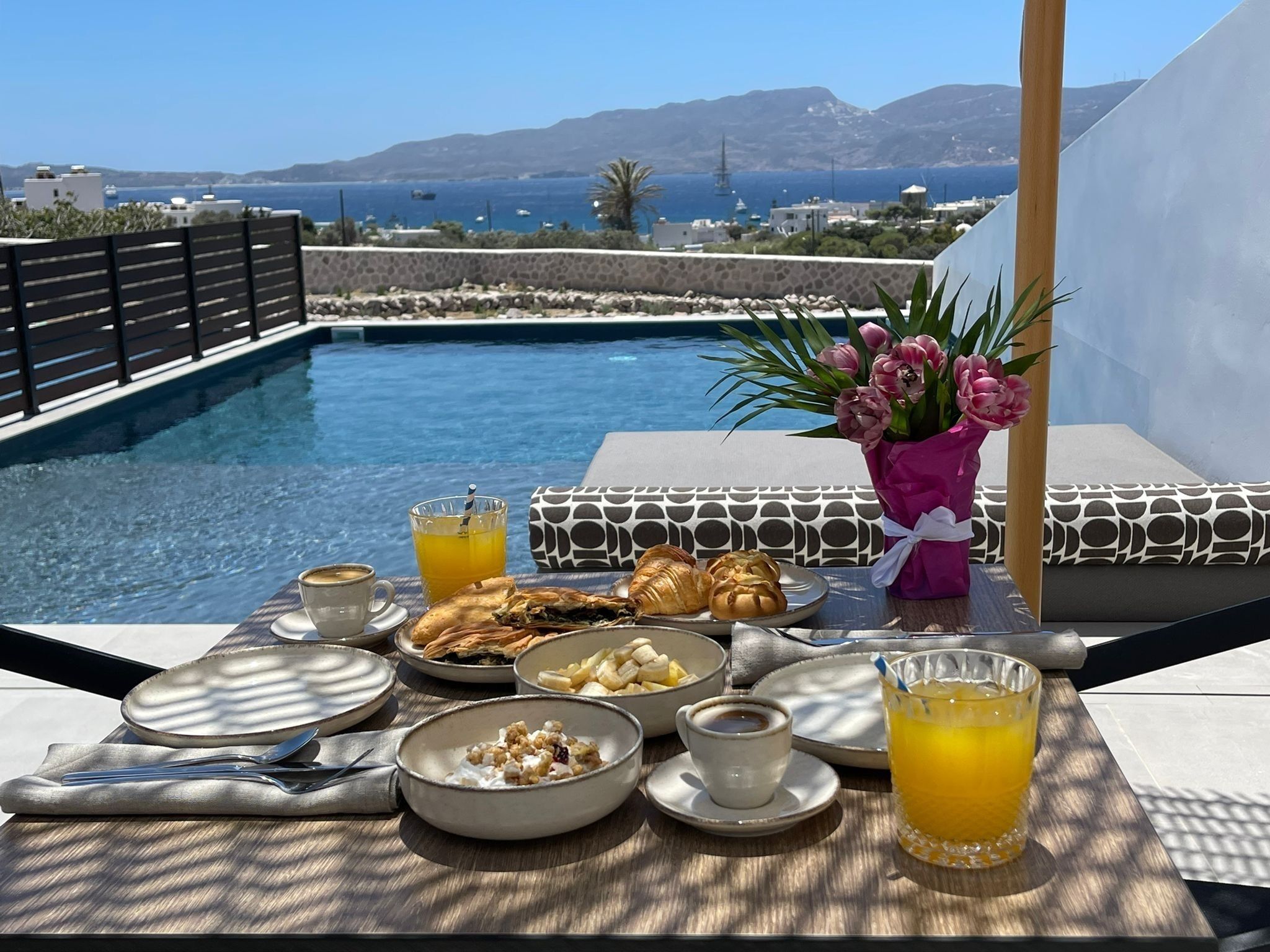 A dining table with breakfast items, a vase of flowers, and a view of a pool and sea.