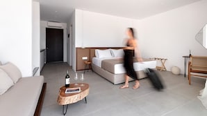 Une chambre d’hôtel moderne avec un canapé, un lit, une table basse ronde en bois sur laquelle se trouvent une bouteille et des verres, et une personne qui marche.