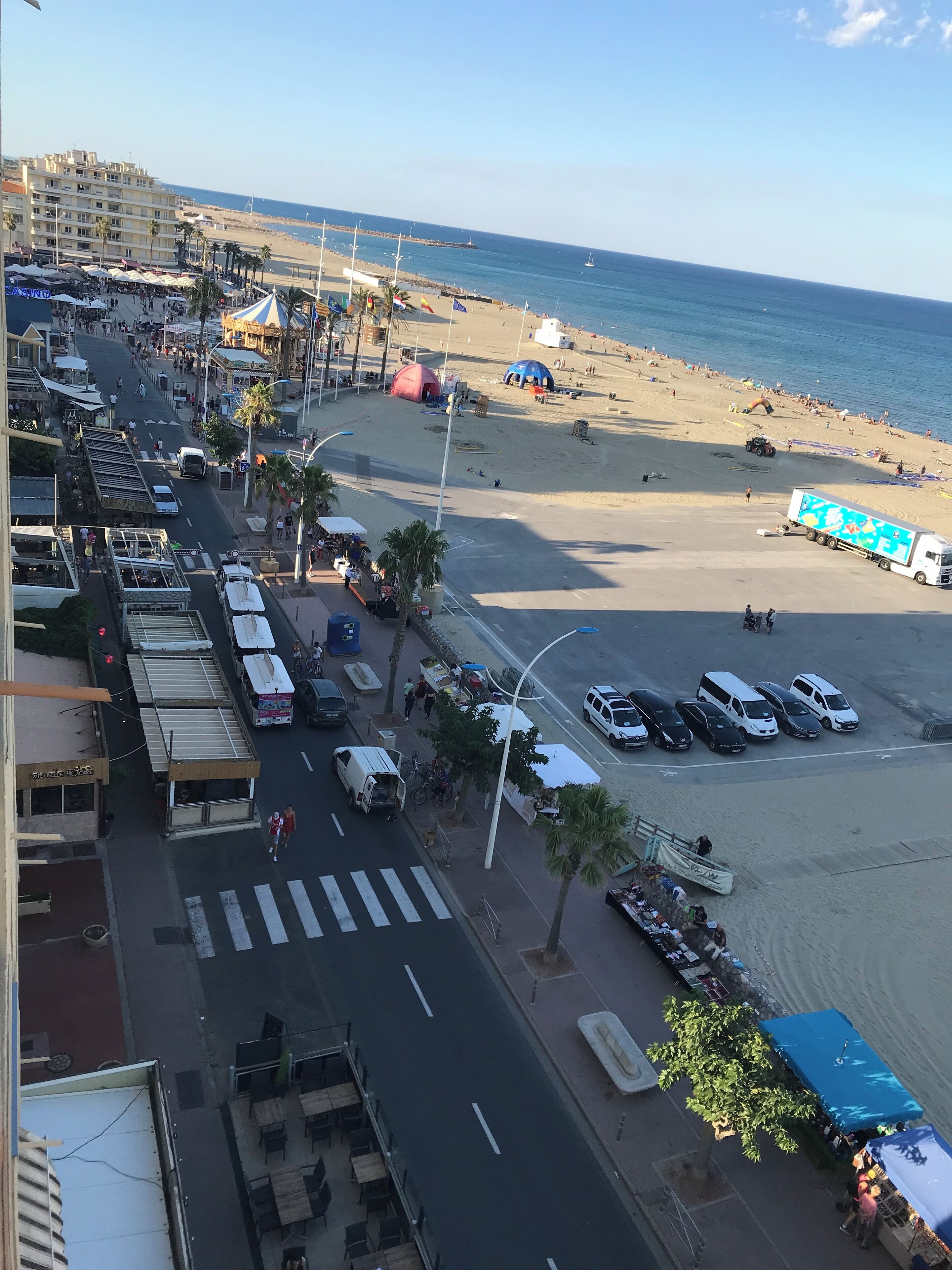 A coastal street with a beach in the background, featuring a pedestrian crossing, parked vehicles, and outdoor seating areas.