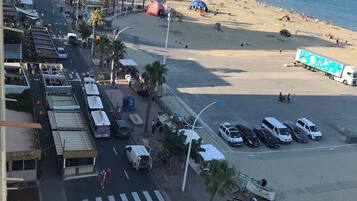 A coastal street with a beach in the background, featuring a pedestrian crossing, parked vehicles, and outdoor seating areas.