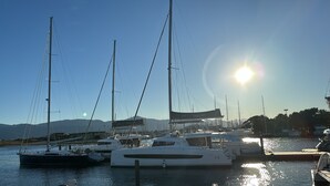 A marina with multiple sailboats docked, a clear sky, and mountains in the background.