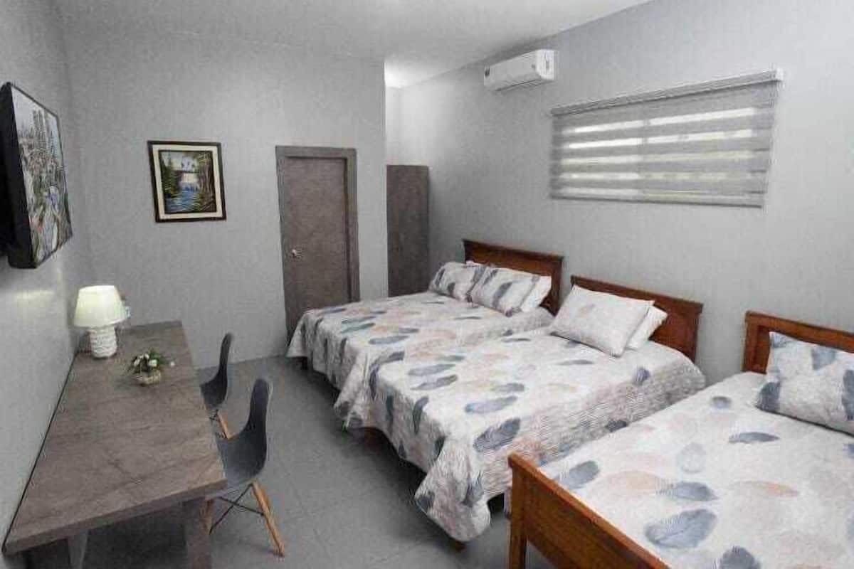 Quarto com duas camas, uma escrivaninha, uma cadeira, ar-condicionado fixado na parede e janela com persianas.