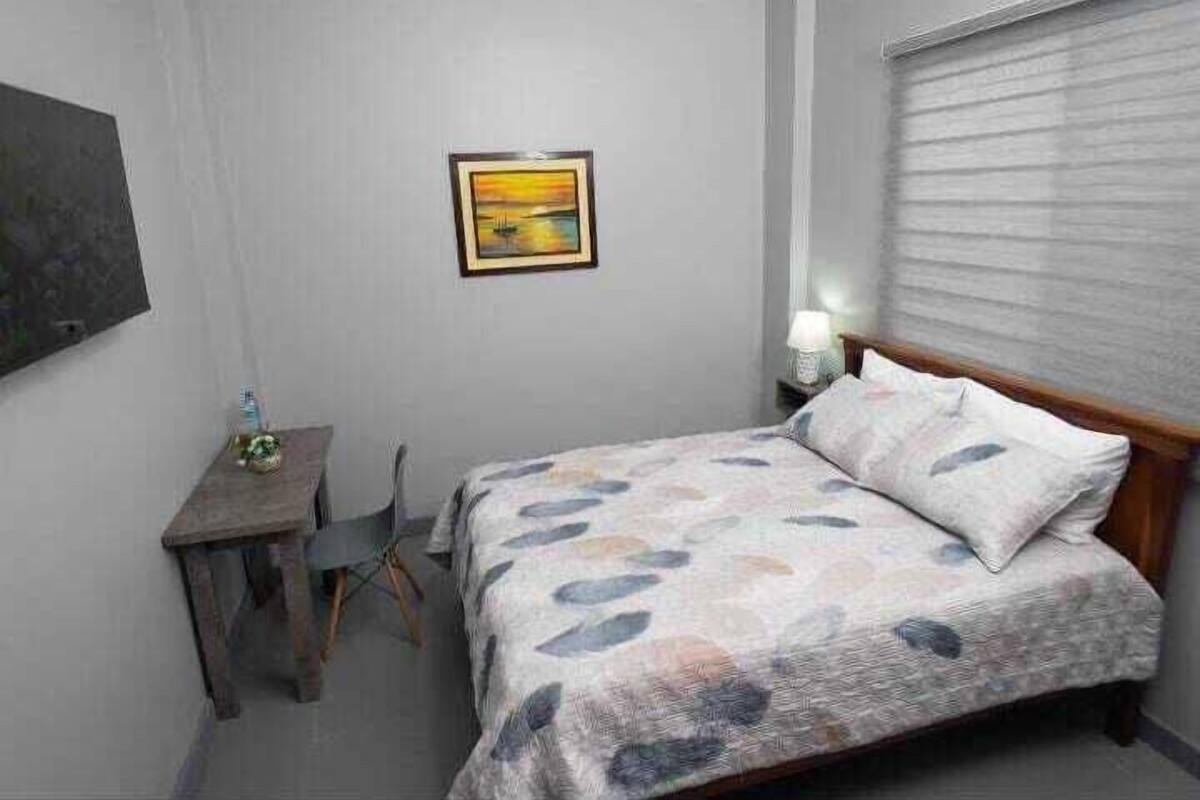 Um quarto pequeno e bem arrumado, com uma cama, uma mesa de cabeceira, uma cadeira e um quadro na parede.