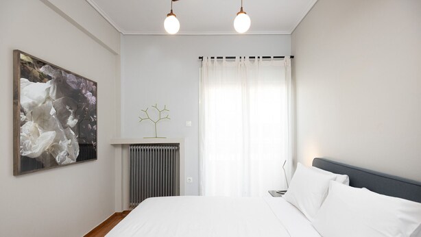 A bedroom with a bed, a radiator, a wall-mounted light fixture, a window with sheer curtains, and a framed picture on the wall. - Stylish 2 bedroom Apt in Thiseion, large balcony (Athina)