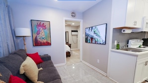 A modern living room with a grey sofa, red and beige cushions, a painting of a rooster, and a kitchen with white cabinets and a tiled backsplash. - Location, Location, Location! Welcome to a charming 1BR in Uptown New Orleans.  (New Orleans)