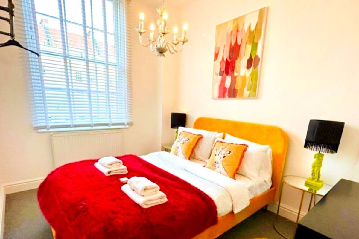A bedroom with a bed, a red blanket, a chandelier, a painting, and a nightstand with a lamp.