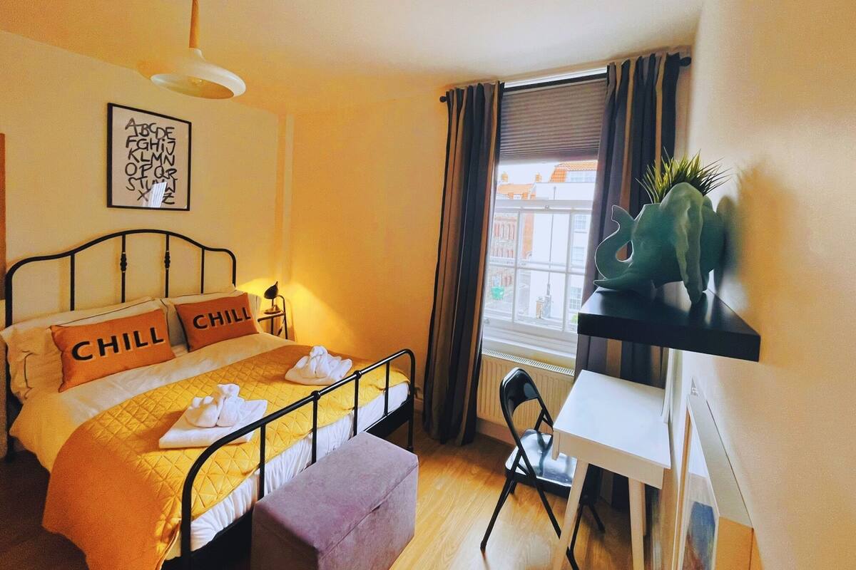 A bedroom with a bed, pillows, a desk, a chair, a window, and a plant.