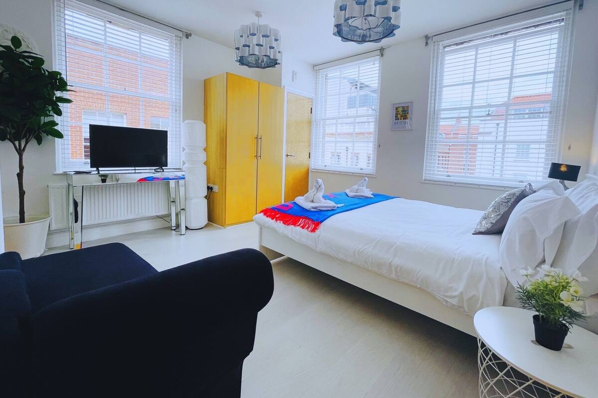 A modern hotel room with a bed, a desk with a computer, a yellow wardrobe, and large windows.