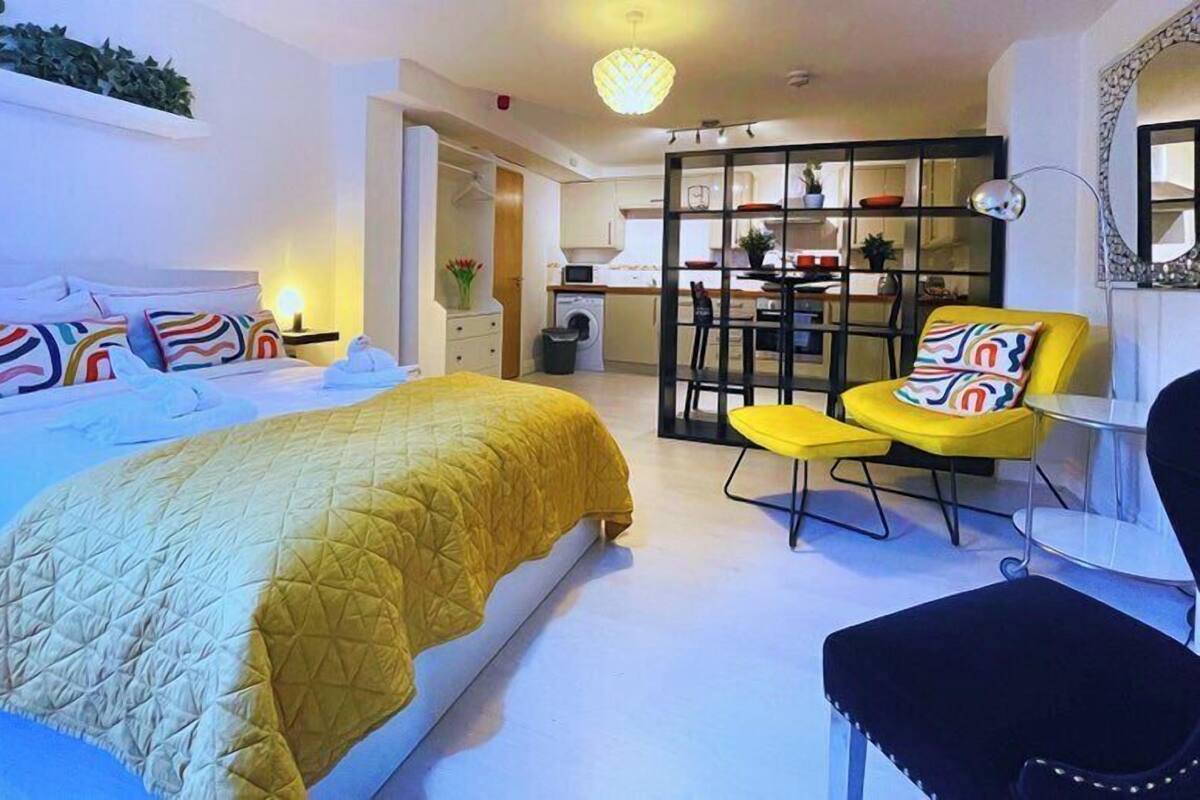 A modern hotel room with a large bed, a yellow chair, a small table, and a kitchenette in the background.