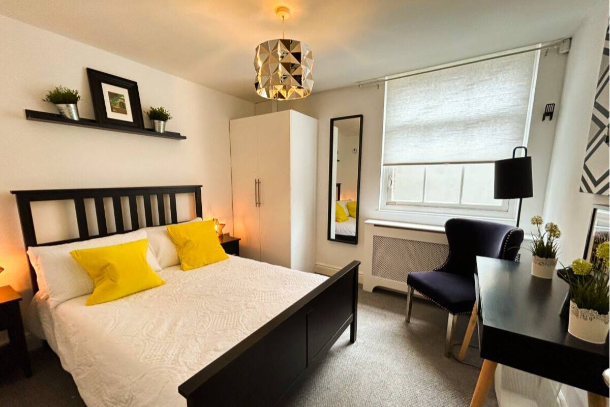A bedroom with a bed, bedside table, chair, desk, and a window with blinds.