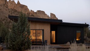 A modern house with a wooden exterior, a large window, and a door leading to an interior with a sofa and a chair.