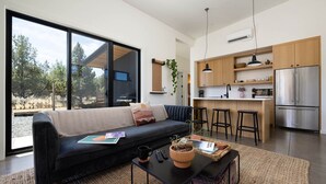 A modern living room with a black sofa, a coffee table, and a kitchen area with wooden cabinets and a stainless steel refrigerator.