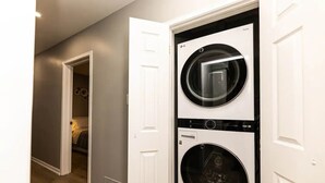 Ein Waschraum mit zwei übereinander gestapelten Frontlader-Waschmaschinen und Trocknern, einem Holzboden und einer Tür zu einem Schlafzimmer.