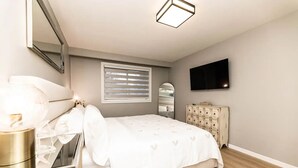 Ein Schlafzimmer mit Bett, Nachttisch, Kommode, Spiegel, Fenster und einem Flachbildfernseher.