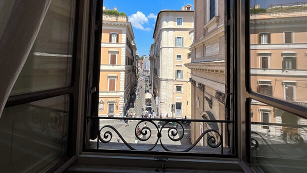 Vista desde una ventana con barandillas de hierro forjado, sobre una calle con edificios históricos y un cielo azul despejado.